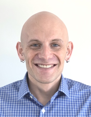 A headshot of a smiling bald person with earrings, in a blue and white checked shirt, against a plain pale background.