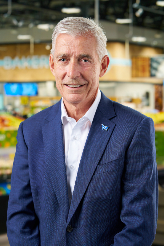 Greg Foran, chief executive of Kroger, smiling in a blue jacket and an open-collared white shirt, in front of a blurred grocery store bakery.