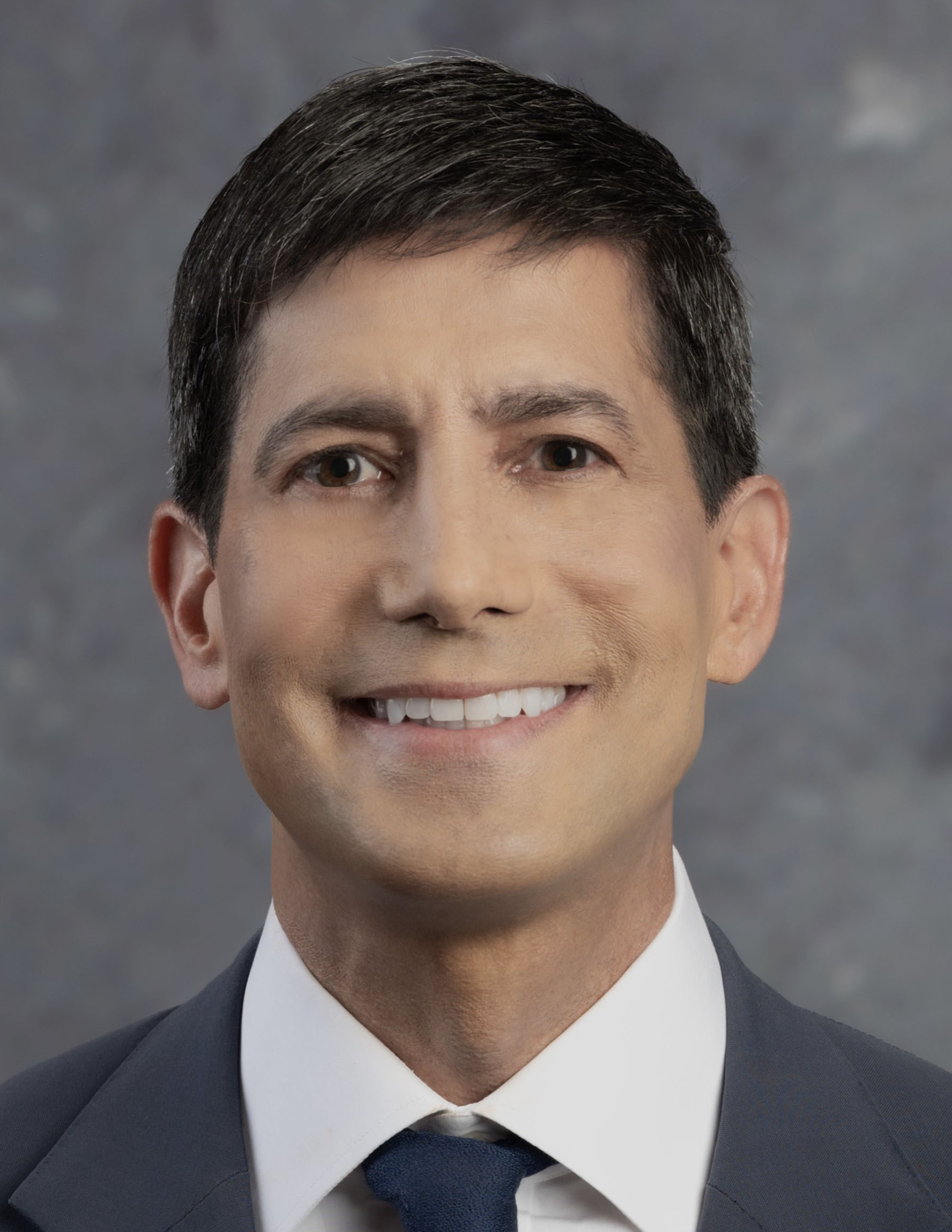 A studio headshot of a smiling person with short dark hair, in a charcoal suit, white shirt and navy tie, against a mottled grey backdrop.