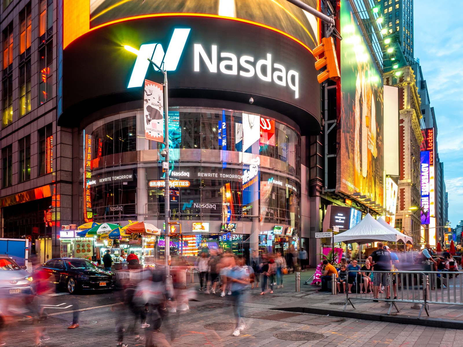 Nasdaq's tower in Times Square at dusk: a curved building wrapped in screens, with a huge lit Nasdaq sign and logo across its dark top band. Below it, a glass front with more screens, street-food carts under striped umbrellas, parked cars and a blurred crowd crossing the street, with bright billboards on the buildings to the right.