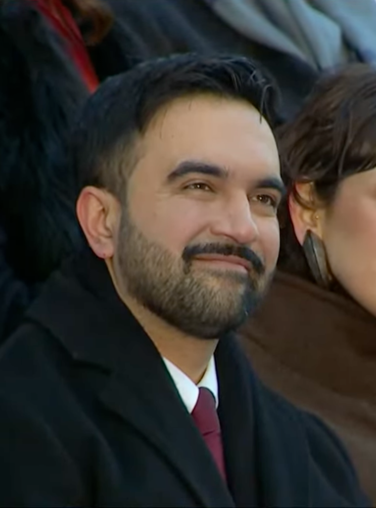 Zohran Mamdani in a dark overcoat, white shirt and maroon tie, smiling slightly, with another person out of focus behind his shoulder.